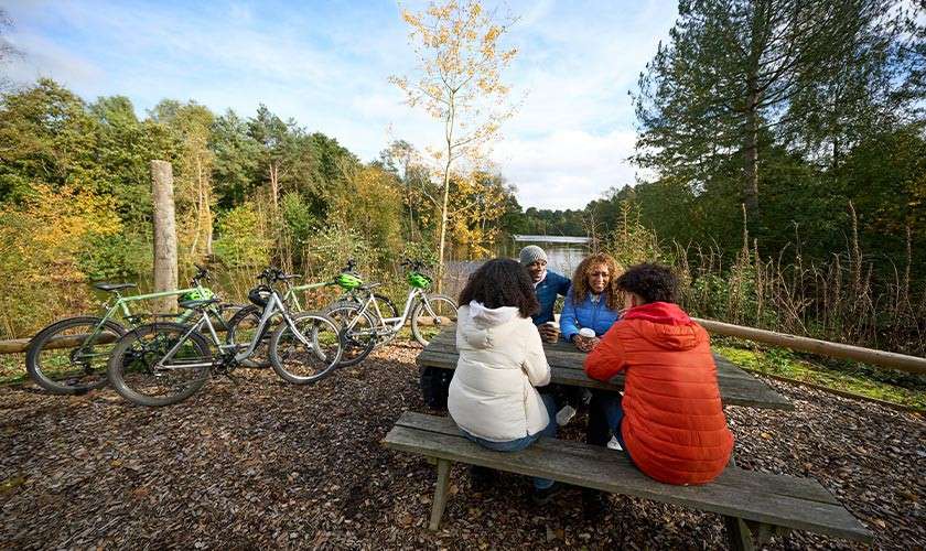 Four cyclists sit and chat at a wooden picnic table, sipping drinks, while their bikes lean against a rail in a forest clearing under sky during a rest stop.
