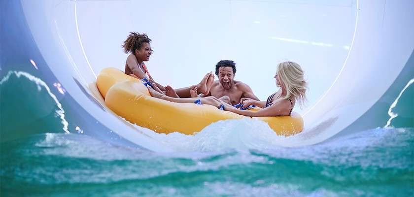 Three riders grip a large yellow inflatable raft, splashing and screaming as it descends a curved, enclosed water slide, surrounded by turquoise water and white tunnel walls.