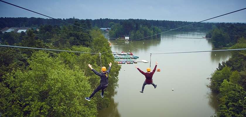 Two harnessed riders zip-line with arms outstretched over a calm lake, above docks and moored boats, surrounded by dense forest under a cloudy sky; cables converge toward a distant platform.