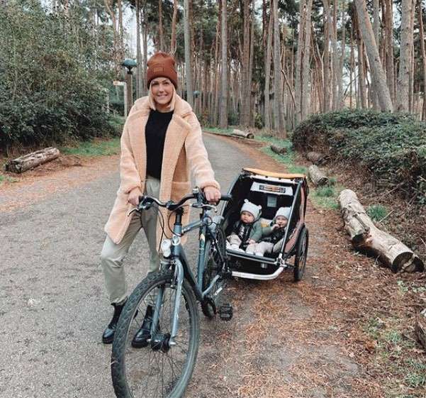 A woman standing by her bike with her two infants in the trailer behind her