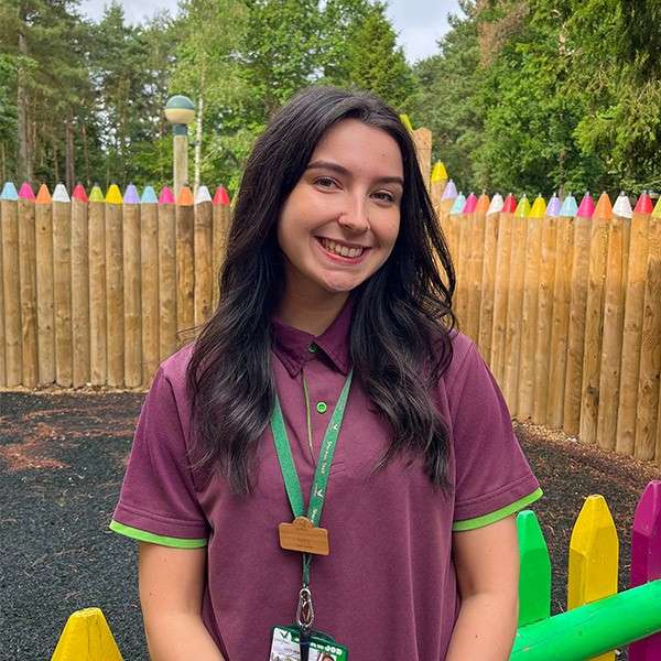 Woman smiles, standing in a purple polo with a lanyard; behind her, a playground with a colorful pencil-shaped fence, dark rubber ground, and trees.