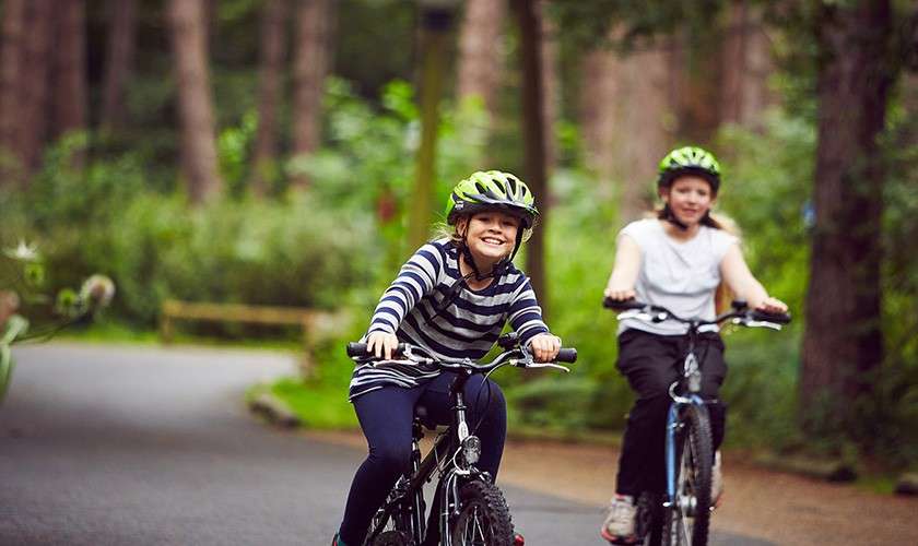 Two children on bicycles ride forward, smiling, wearing green helmets, along a curving paved path through a lush, wooded park with tall trees and soft, dappled light.