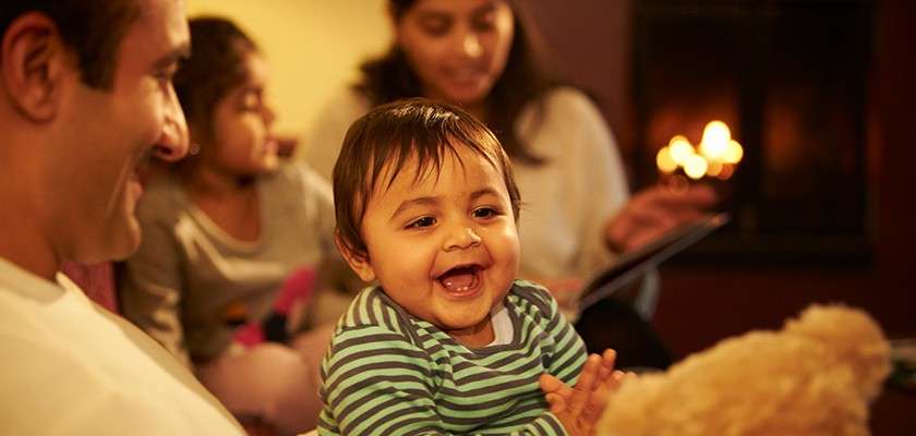 Baby laughs and claps, reaching toward a teddy bear, while seated on an adult’s lap; in the background, two people relax near a glowing fireplace in a warmly lit room.
