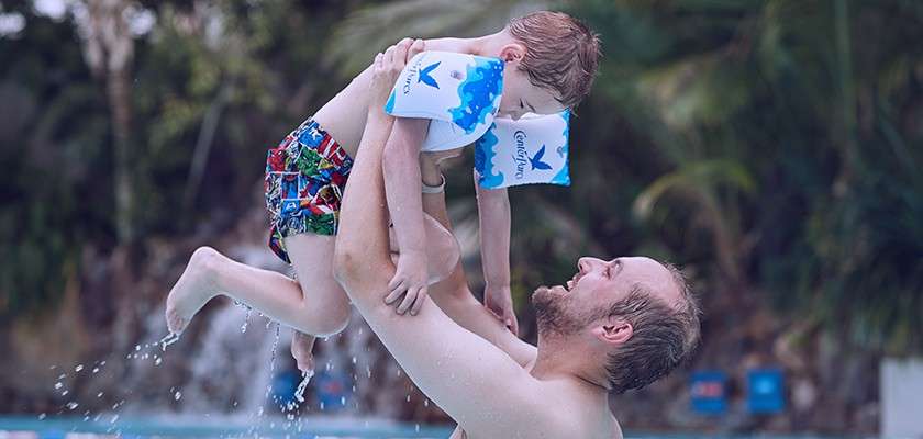 Child wearing inflatable armbands is lifted playfully by an adult in a swimming pool, water dripping, surrounded by lush tropical plants in the background.