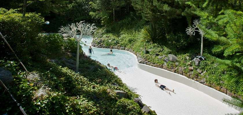 Children and adults float and slide through a shallow, curving water channel, some wading. Set amid dense greenery and rocks, with white tree-like spray sculptures lining the banks.