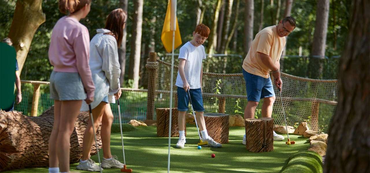 Family group plays mini-golf, swinging putters toward colorful balls on artificial turf, while others watch. Surrounded by trees, wooden stump obstacles, rope fencing, and a yellow flag under forest light.