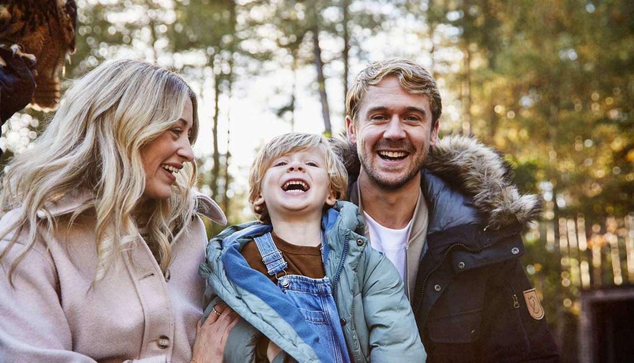 Three people laugh together; the child in a blue overall is held between two adults, all wearing winter coats; background shows sunlit trees in a forest or park.