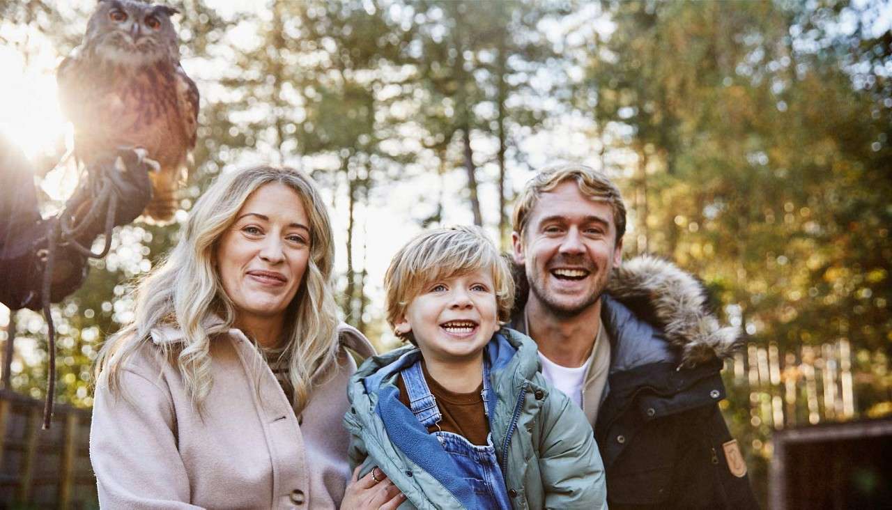 Family of three smiles at the camera, the child centered, while an owl perches on a gloved handler’s arm nearby; background shows sunlit trees in a wooded park.