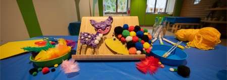 Craft supplies sit assembled on a blue-covered table: pom‑poms, feathers, felt shapes, yellow yarn, tweezers, and a wooden tray with an animal cutout, in a classroom with windows and stools.