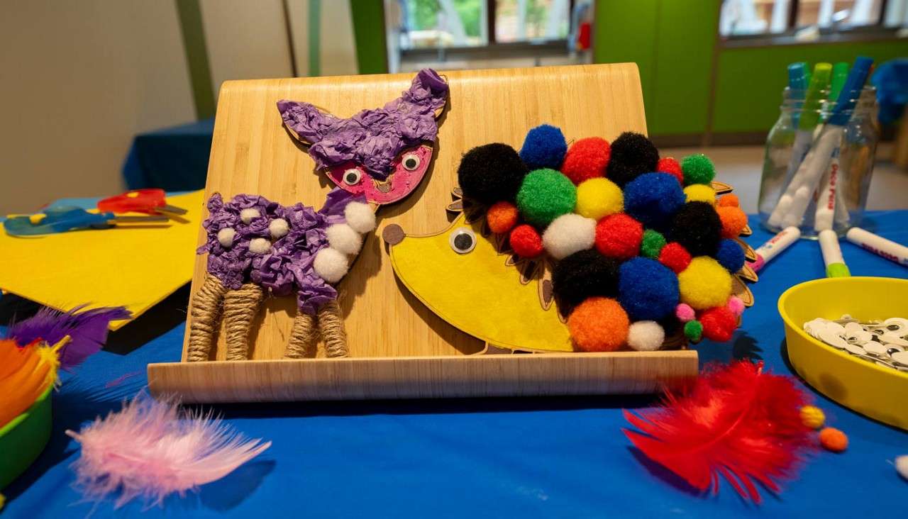 Two kid-made animal collages sit on a wooden stand: a purple fox and a yellow hedgehog with pom‑pom spines, surrounded by craft supplies—feathers, googly eyes, markers, and paper—on a classroom table.
