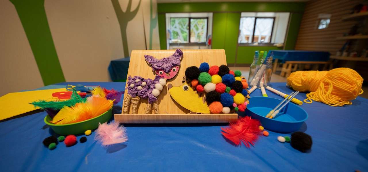 Animal crafts—a purple creature and a pom-pom-covered hedgehog—stand on a wooden holder, surrounded by yarn, markers, feathers, and bowls of supplies on a blue table in a classroom.