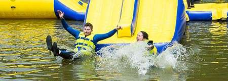 Two people wearing yellow life vests slide down a bright inflatable into the water, splashing and holding hands, within a floating blue-and-yellow water park on a lake.