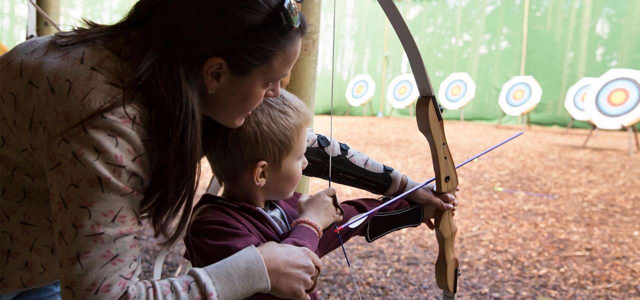 Child archer draws a bow as an adult guides their hands, both wearing armguards; they aim an arrow toward circular target boards in an indoor-outdoor archery range with woodchip ground.