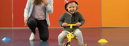 Toddler on a small yellow bike rides forward, smiling, wearing a red helmet. Nearby, an adult kneels supervising. Context: indoor gym floor with colored wall panels and scattered training cones.