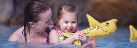 Child grips a yellow inflatable shark while smiling; adult supports the child. They wade in shallow pool water, splashing gently, with a blurred rocky backdrop suggesting an indoor aquatic setting.