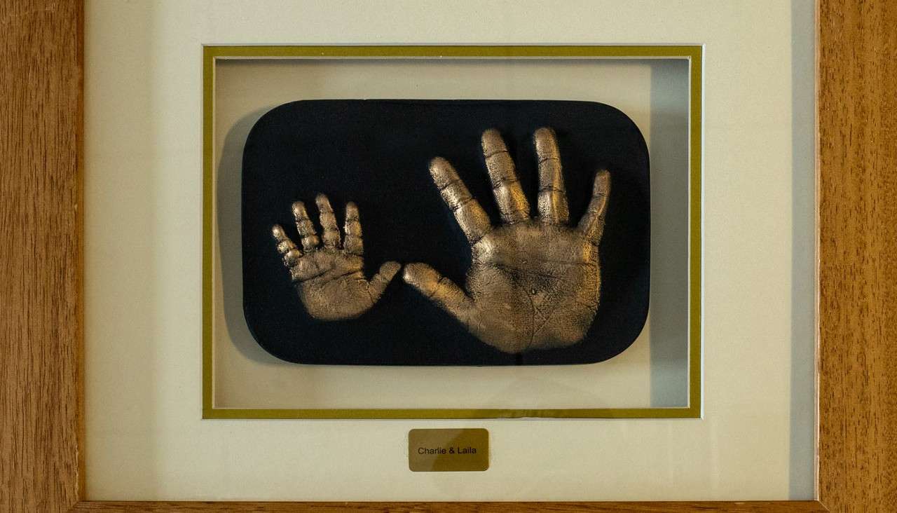 Two metallic handprint casts—one small, one large—rest side-by-side on a black plaque. They are mounted in a matted wooden frame with a brass nameplate reading: Charlie & Laila.