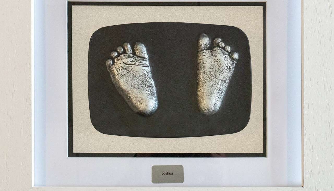 Silver baby footprints—two cast soles—sit mounted on a black oval, displayed in a white frame on a wall, with a small plaque reading: Joshua.