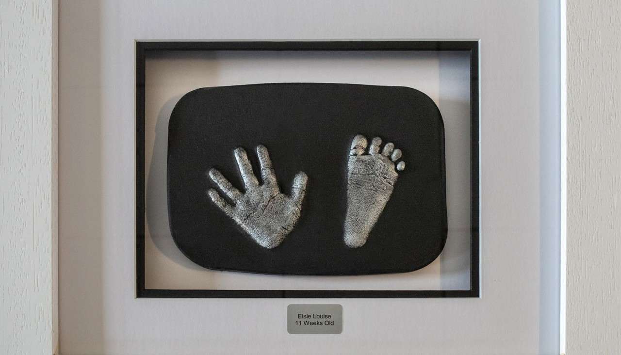 Framed keepsake casts display a baby's silver handprint and footprint, mounted on a black plaque within a white shadow box. Text: “Elsie Louise 11 Weeks Old”.