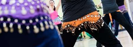 Dancers’ hips sway, jingling coin-adorned scarves; in a bright studio, participants in black pants mirror movements, legs bent and arms active, suggesting a belly-dance style class.