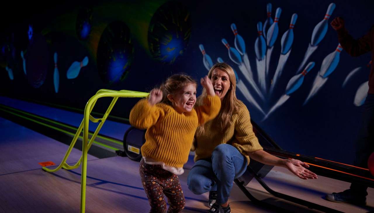 Child jumps and cheers, adult kneels clapping, after rolling a ball. Context: neon-lit bowling alley with a green ramp, ball return, and wall mural of flying pins and planets.