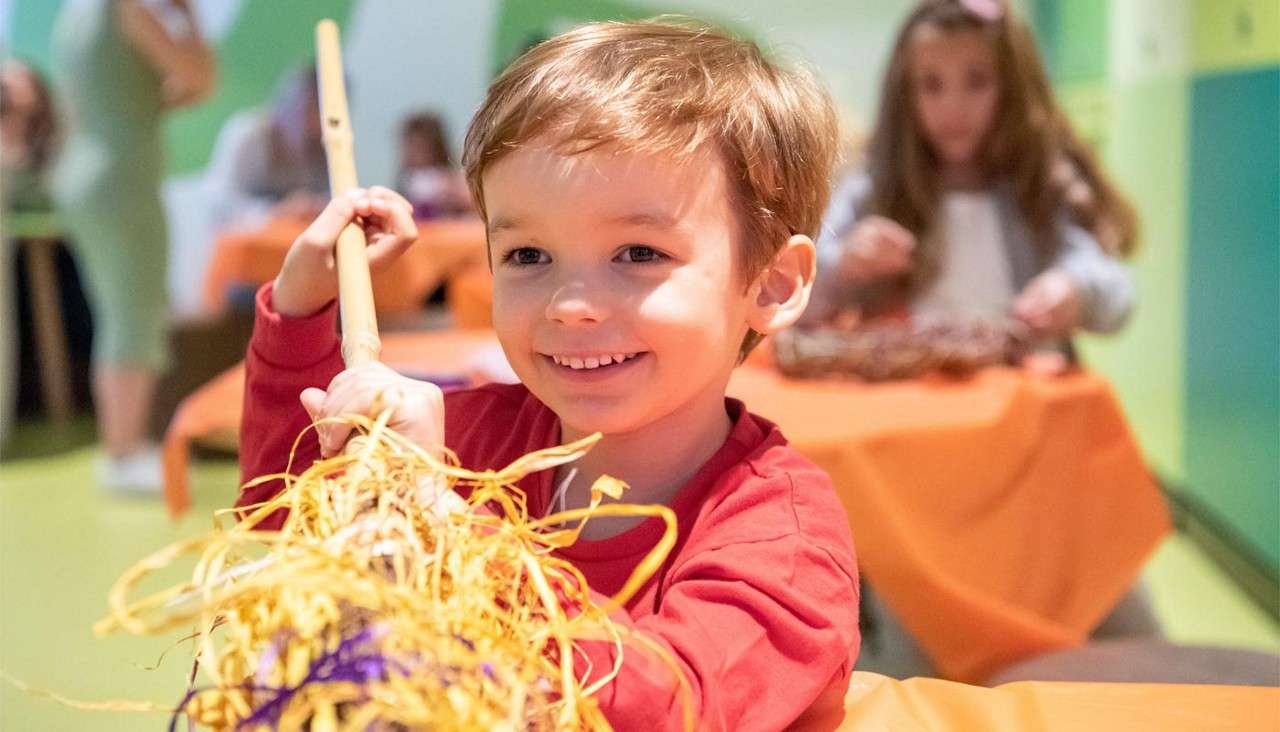 Smiling child holds a stick wrapped in yellow and purple raffia, crafting at a table, with other children and orange-covered tables in a bright classroom workshop.