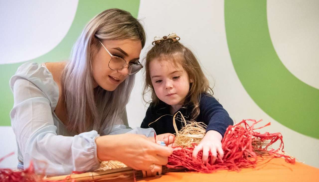 Two people craft together; a woman guides a young girl threading red raffia through straw on an orange table, concentrating closely. Background shows a white wall with large green curves.