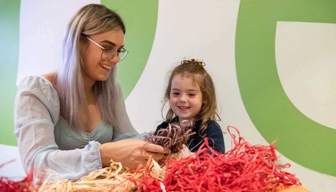 Two people collaborate, sorting colorful shredded paper while smiling. They sit at a table in a bright room with large green wall graphics, engaging in a hands-on craft activity.
