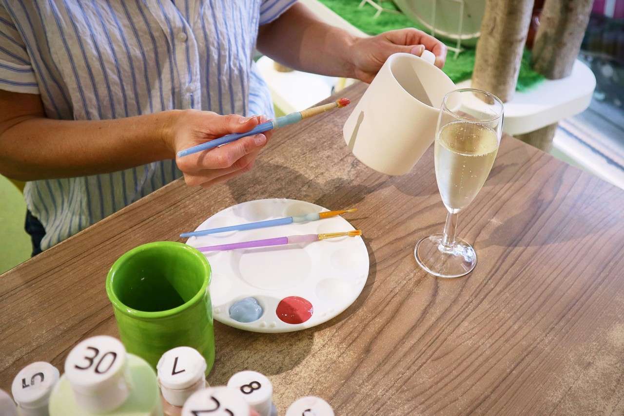 shot of a womans hands painting a mug, with a glass of prosecco on the table next to her