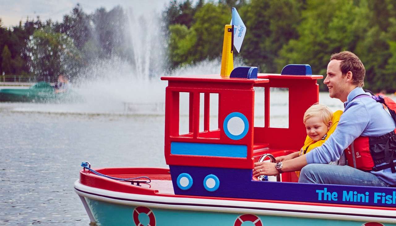 A man and child ride a colorful mini tugboat on a lake, steering together; fountains spray in the background; text on the hull reads: "The Mini Fish".