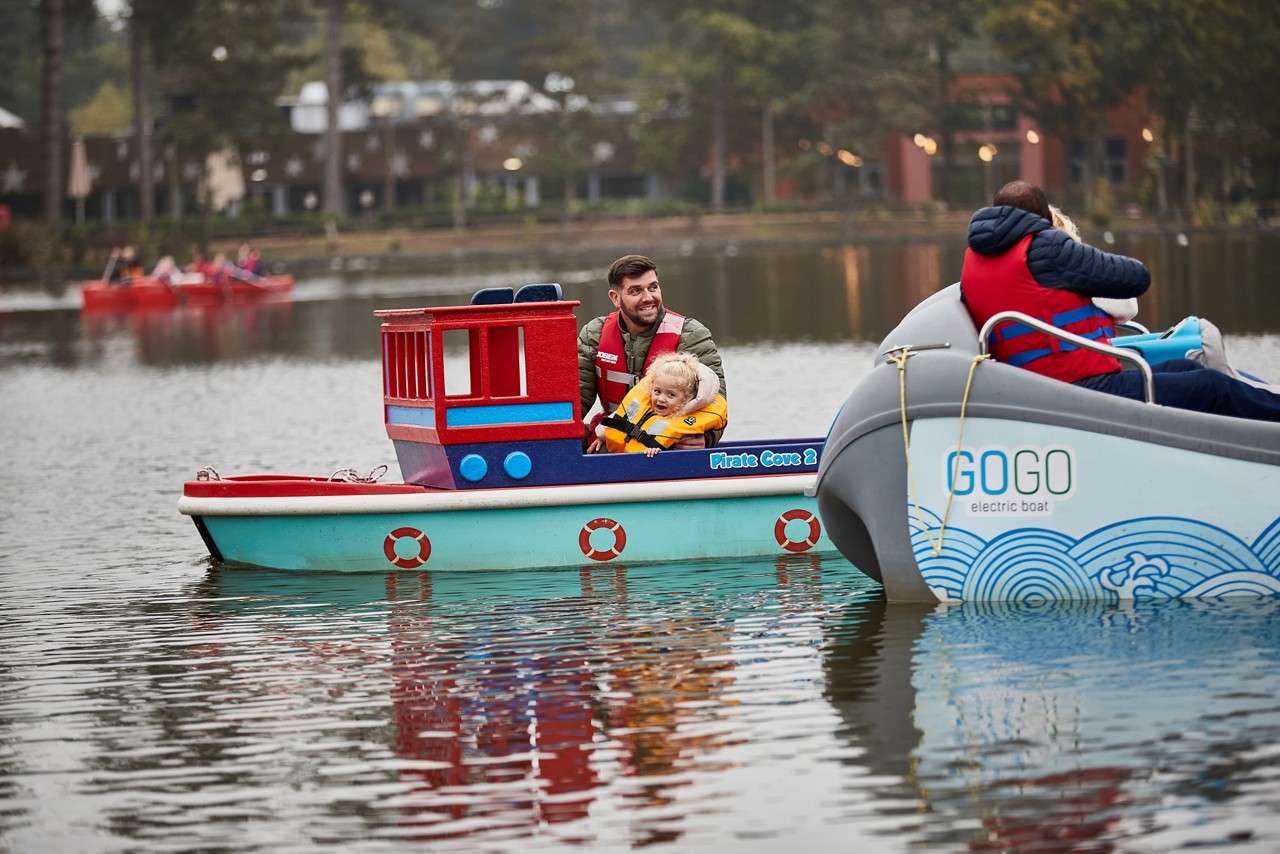 Child wearing a yellow life jacket steers a colorful toy boat while an adult assists, on a calm lake near trees; boat side text reads: "The Mini Fis".