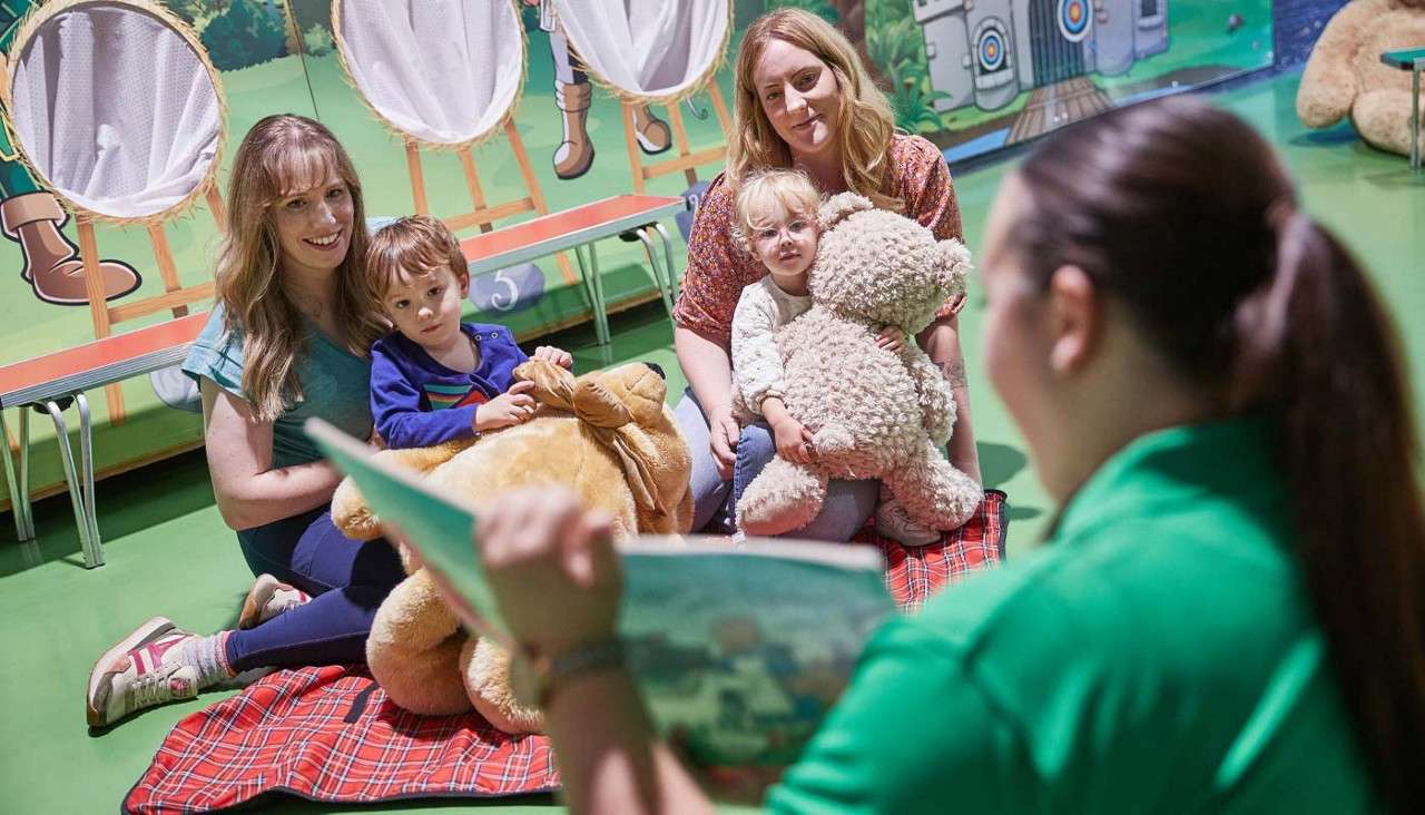 Storyteller reads a picture book to two adults and two children, who cuddle teddy bears, seated on a red blanket in a colorful playroom; bench labeled “5” behind them.