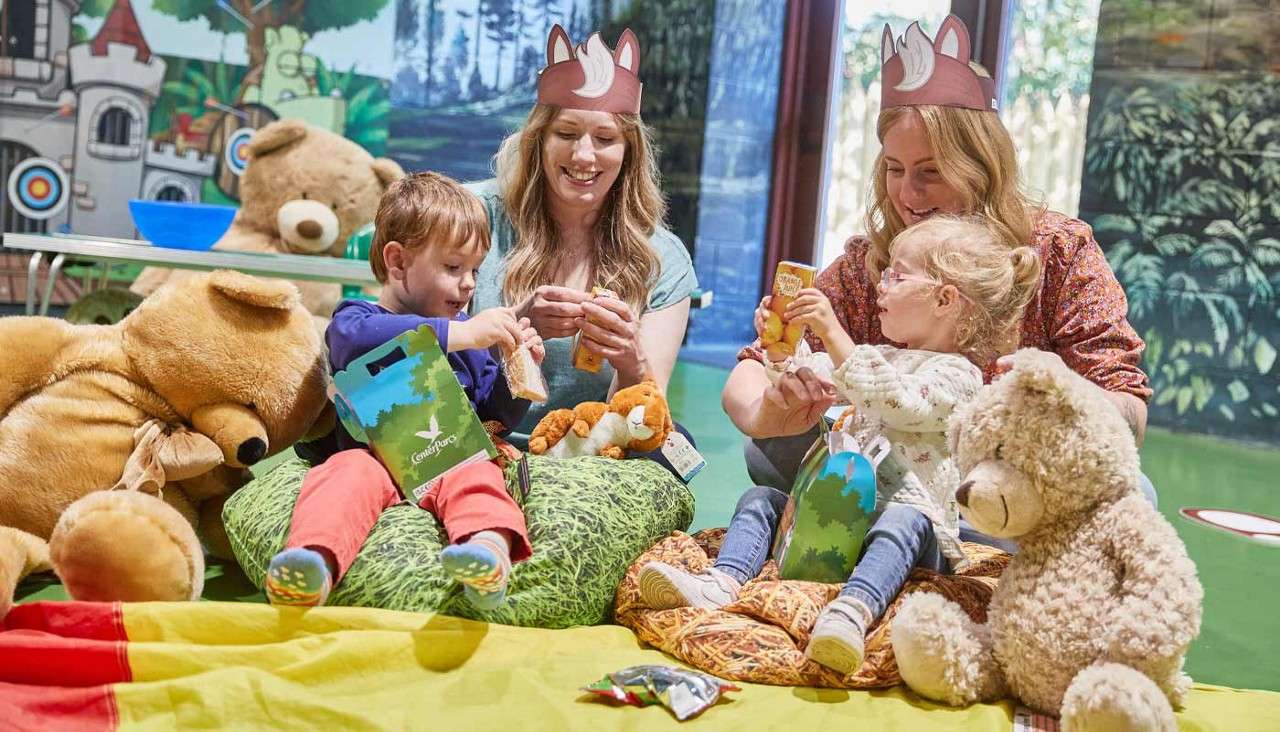 Children and adults wearing fox-ear headbands open snacks and toys while sitting on cushions among teddy bears; a forest-themed mural and picnic blanket surround them. Text: Center Parcs.