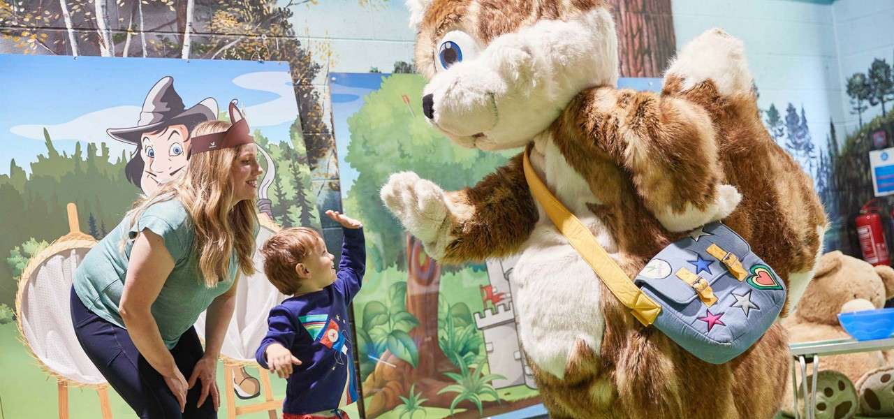 A large squirrel mascot raises a paw to high-five a small child, while an adult smiles nearby in an indoor play area decorated with cartoon forest murals and props.