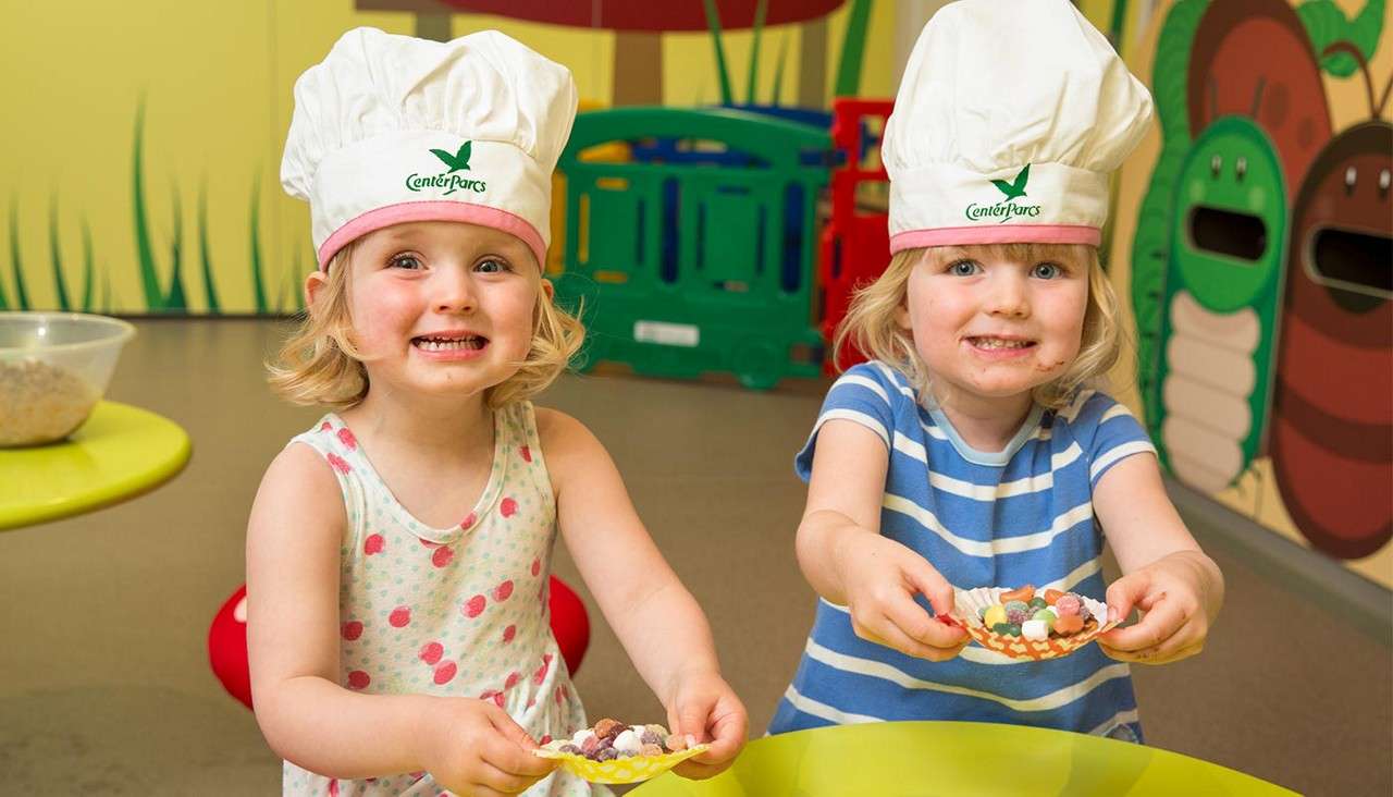 Two toddlers wearing white chef hats hold candy-filled cupcake liners, smiling toward the camera, in a colorful playroom with cartoon bug walls and plastic play structures. Hats read: "CenterParcs" "CenterParcs".