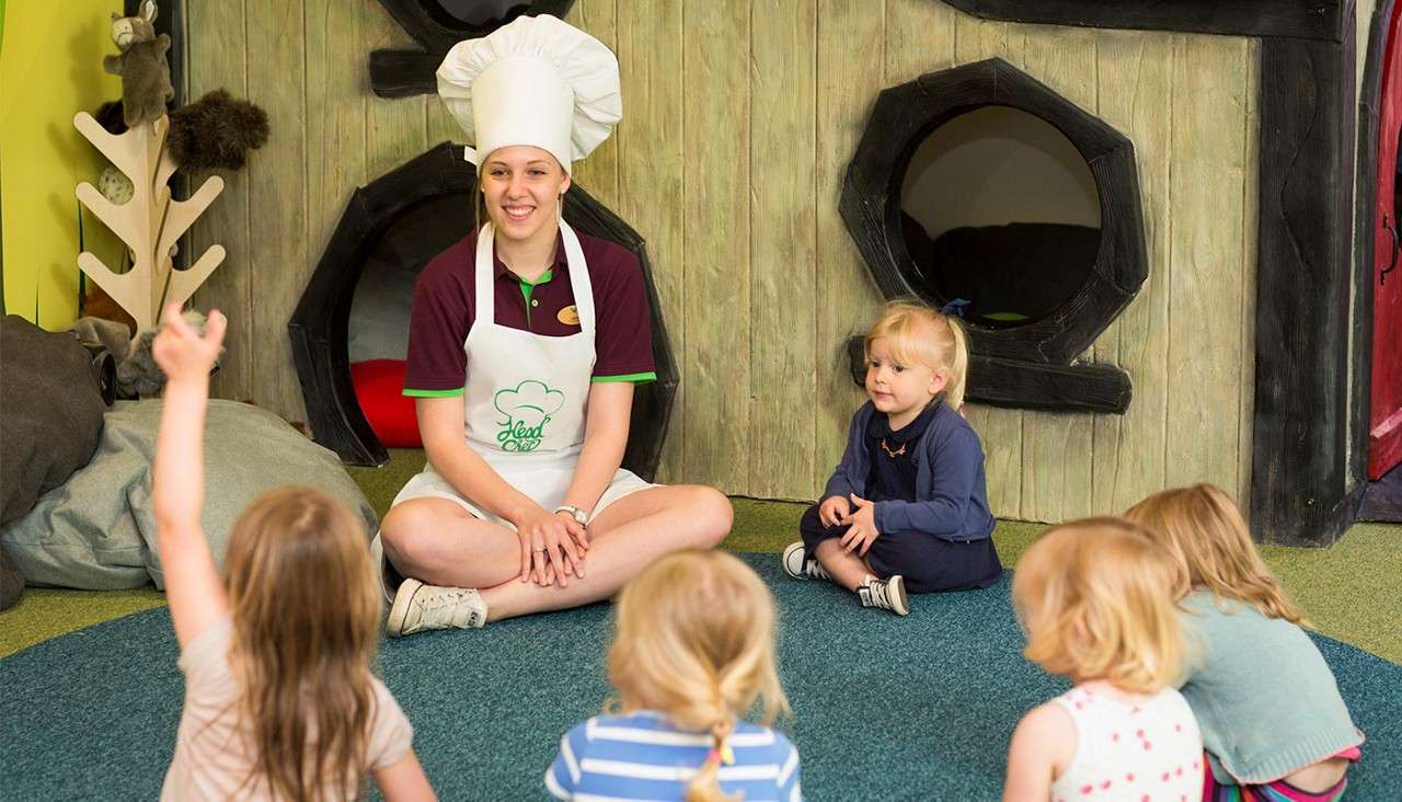 Adult in chef hat and "Head Chef" apron leads storytime, sitting cross-legged while preschoolers sit around, one raising a hand, inside a playroom with faux wooden walls and circular openings.