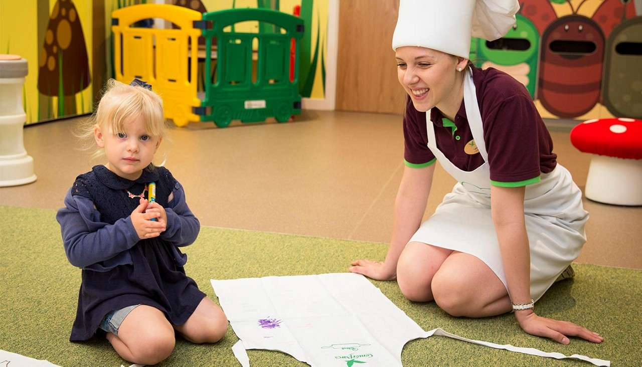 Child holds crayons, drawing on a white apron as a smiling adult in a chef hat kneels nearby; colorful playroom shows cartoon bugs, green fence, and mushroom stool. Text: Center Parcs.