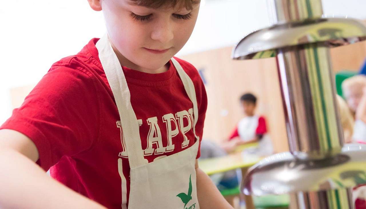 Child in red shirt and white apron concentrates while preparing food beside a tiered metal fountain-like device, in a bright classroom with other children in the background. Text: HAPP