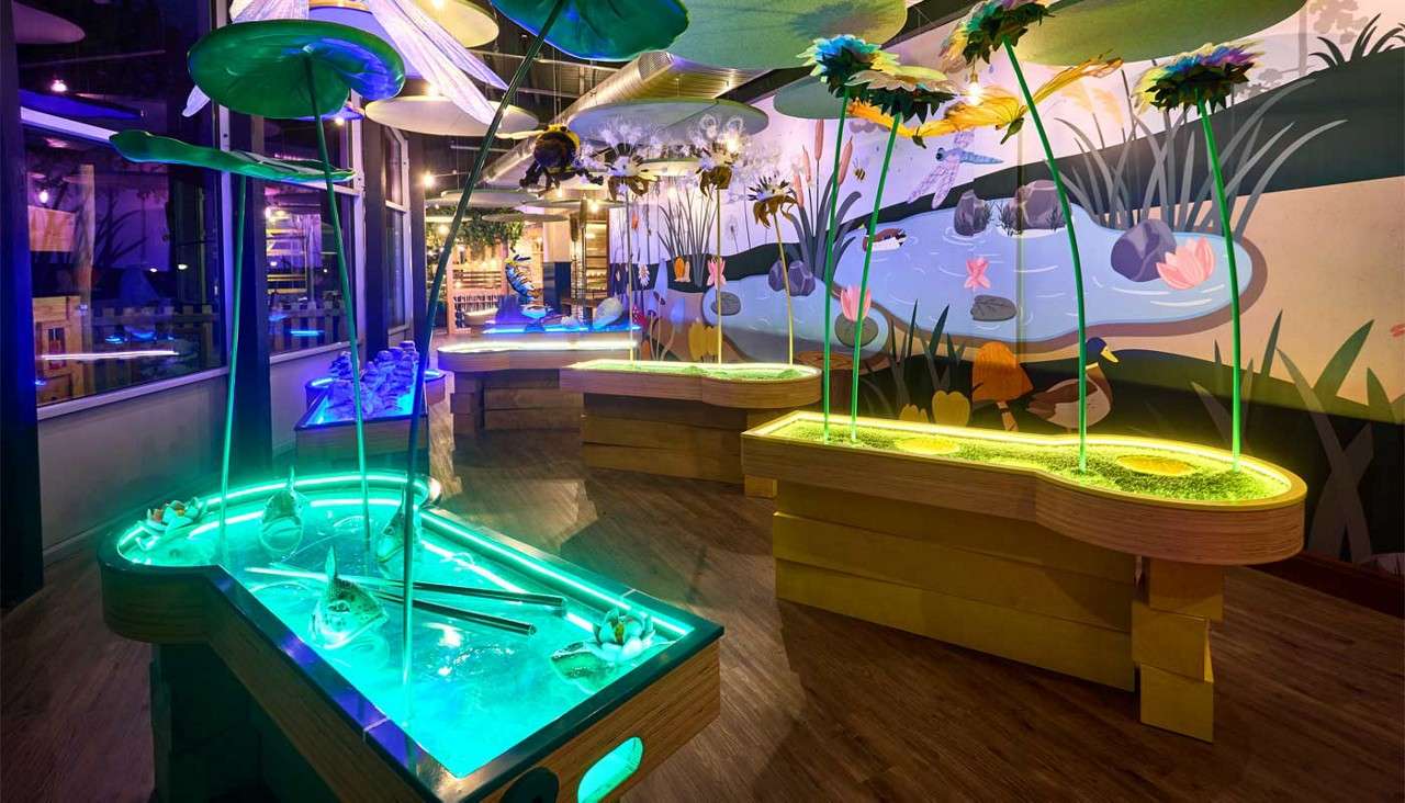 Glowing lily-pad water tables invite hands-on play, casting neon light. Surrounding them, a pond-themed room features hanging leaves, flower sculptures, and a mural of reeds, rocks, ducks, and fish.