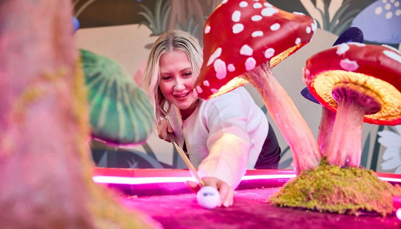 Person aims a cue at a white ball, preparing a shot amid oversized red-and-white mushrooms on a neon-lit, whimsical indoor mini-golf course with mossy bases and a mural-style forest background.