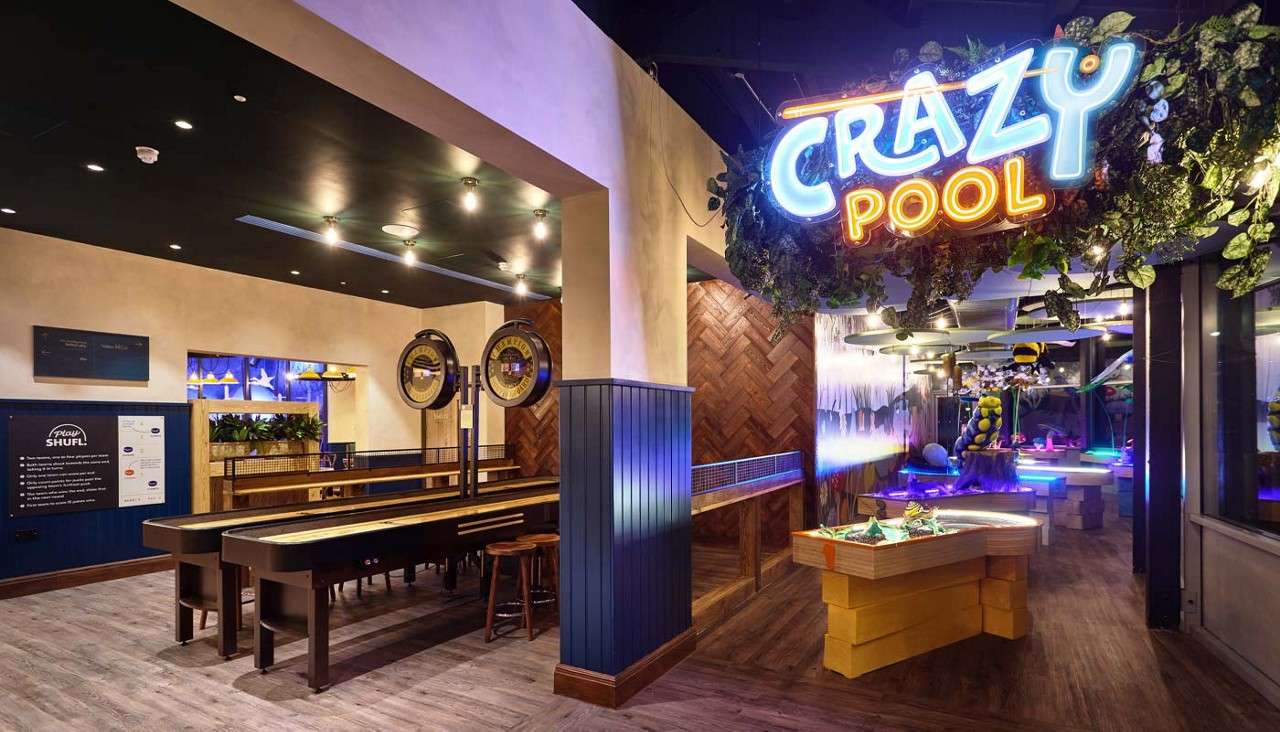 Neon sign glows, advertising CRAZY pool; patrons would play on illuminated mini-golf-style pool tables and long shuffleboard lanes in a modern indoor game bar. Text: CRAZY pool; Play SHUFL.