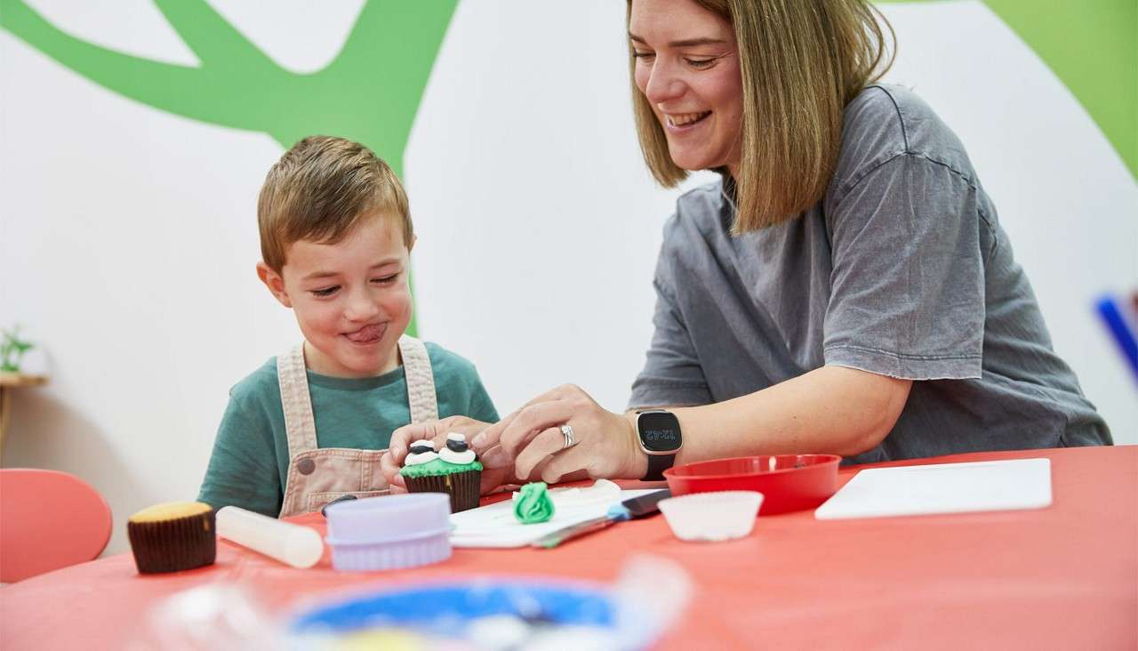 Child and adult decorate a cupcake, applying green frosting and candies, smiling at a red-topped table with bowls, cutters, and utensils, inside a bright room with playful wall graphics.