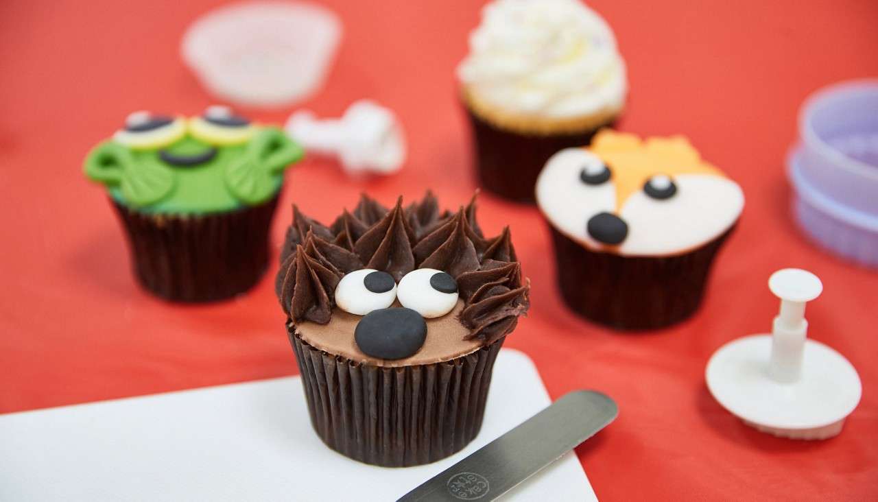 Animal-themed cupcakes sit on a red tabletop, surrounded by decorating tools and a spatula; a hedgehog cupcake is foregrounded, with frog, fox, and whipped-cream styles behind.