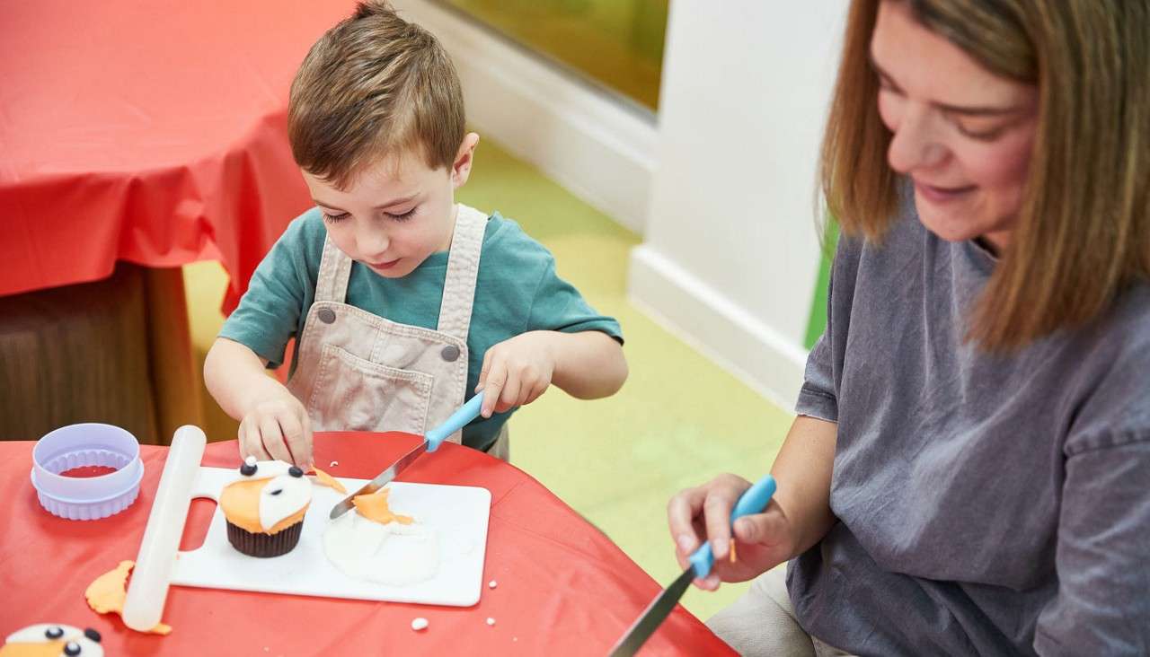 Child decorates a cupcake, spreading orange fondant with a plastic knife, while an adult assists beside them; tools and icing sit on a cutting board on a red-covered table.