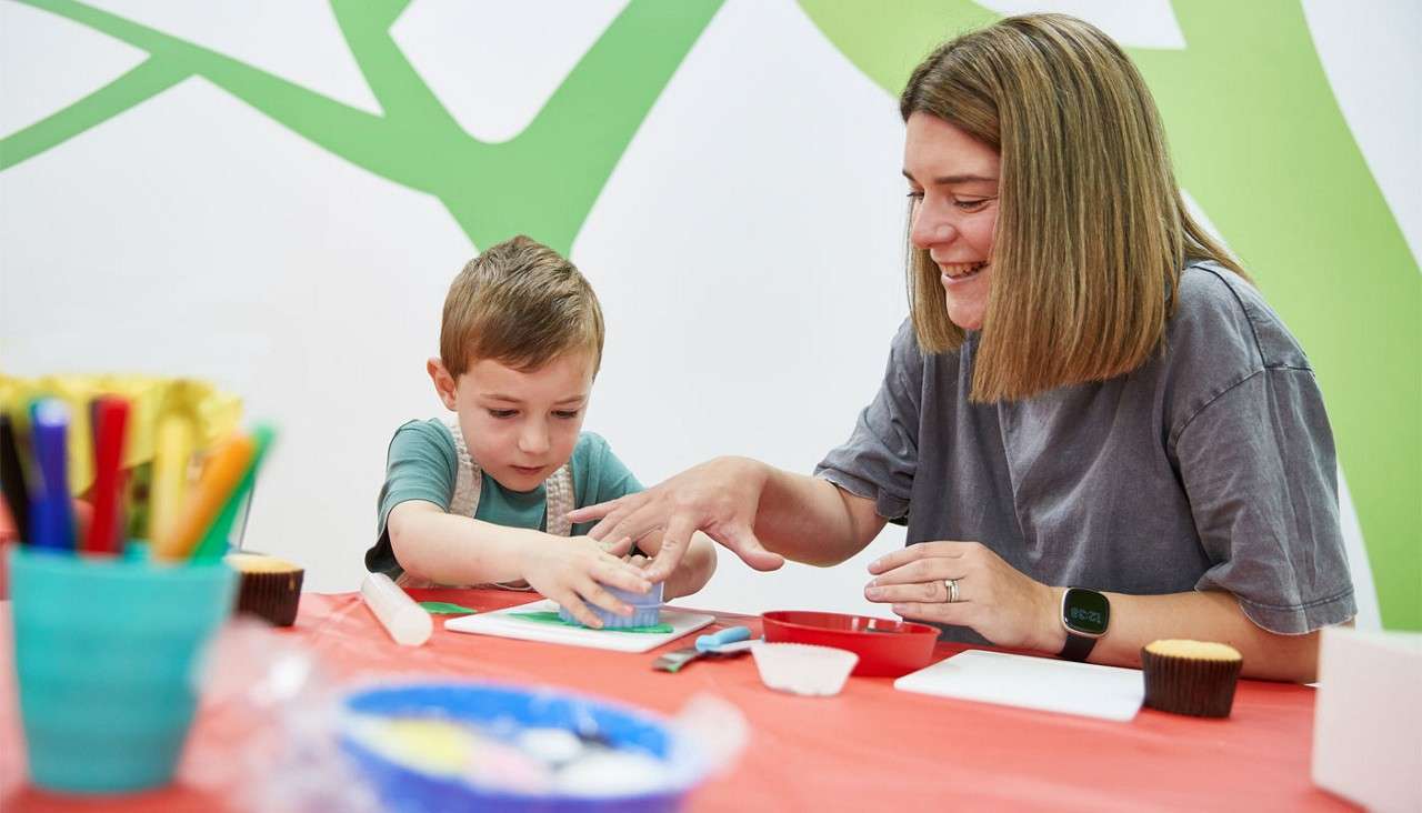 Child presses a round cutter into dough while an adult guides his hand, both seated at a red-covered craft table with utensils, cups, and markers in a bright classroom.