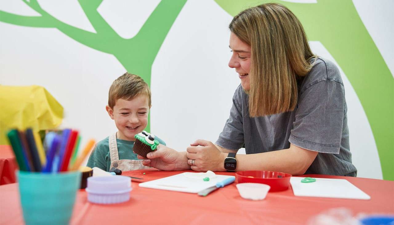 Child presents a decorated cupcake while an adult smiles and guides him, in a bright craft room with a red table, utensils, colored pencils, and a green-and-white wall mural.