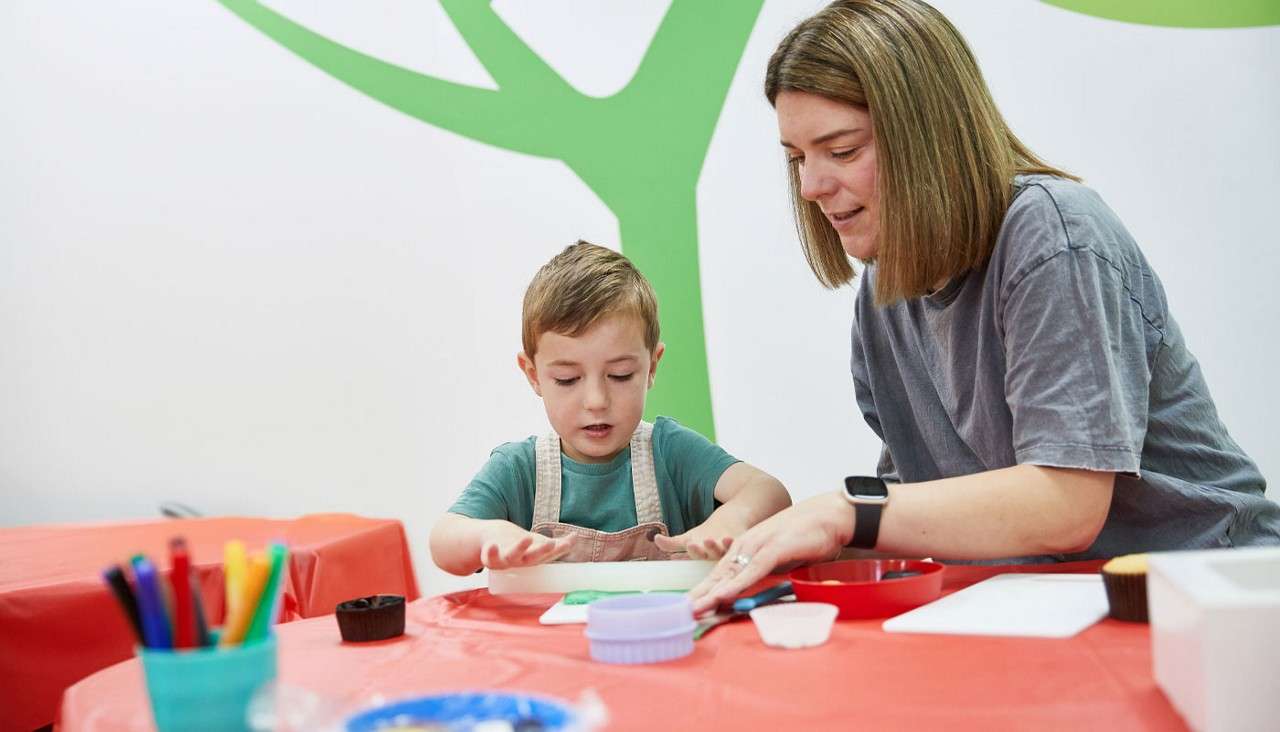 Child presses green dough with hands, adult assists nearby, guiding. In a bright classroom with red tablecloths, art supplies, and cookie cutters on the table, tree mural on the wall.