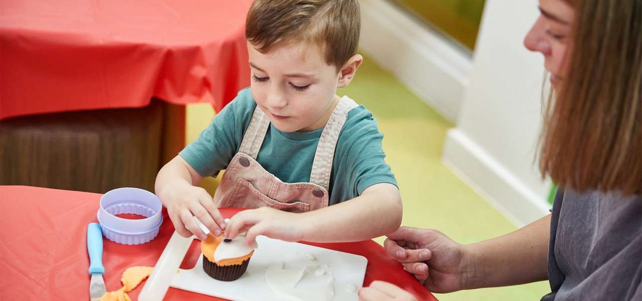 Child decorates a cupcake, pressing orange icing on top while an adult supervises. Plastic cutters and tools rest on a cutting board on a red tablecloth in a bright room.
