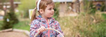 Child holding a wicker basket looks to the side, walking along a garden path; she wears a floral jacket and headband, with blurred trees, grass, and wooden structures in the background.