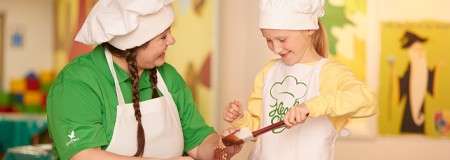 Child and adult chefs scoop chocolate into a measuring cup, collaborating at a table in a bright classroom kitchen with colorful artwork. The child’s apron has printed text that is unreadable.
