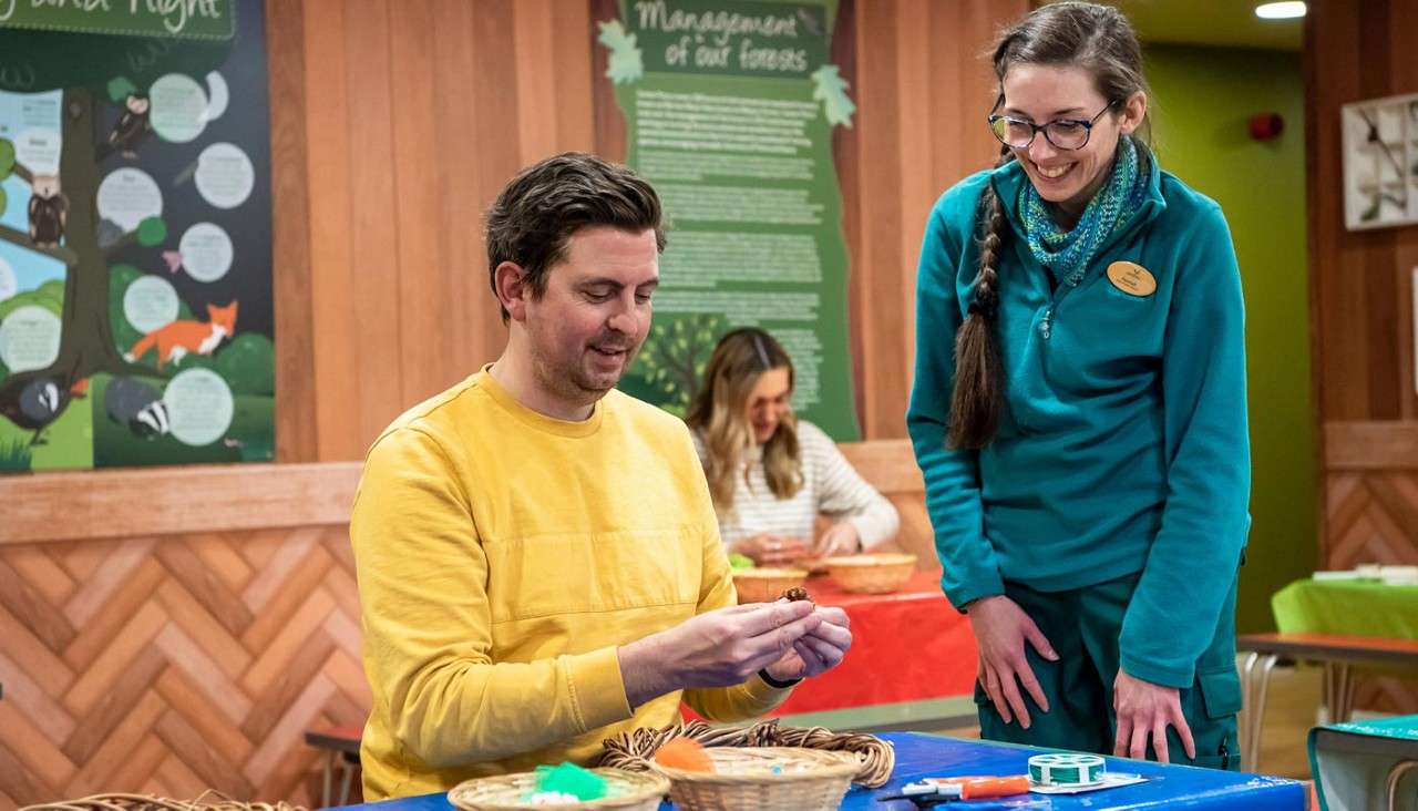 Man crafts a pinecone ornament, observed by a smiling instructor, in a wood-paneled educational room with craft supplies and posters. Visible text: “Management of our forests.”
